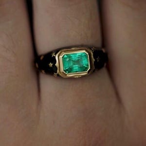 May include: A gold ring with a green emerald gemstone set in a black enamel band. The band is decorated with gold accents.
