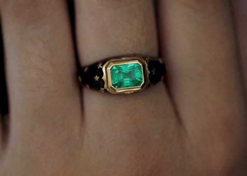 May include: A gold ring with a green emerald gemstone set in a black enamel band. The band is decorated with gold accents.