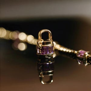 May include: A gold-colored padlock charm with a purple gemstone center, part of a necklace. The charm is rectangular with a curved handle. The necklace chain is also gold-colored, with a blurred background.