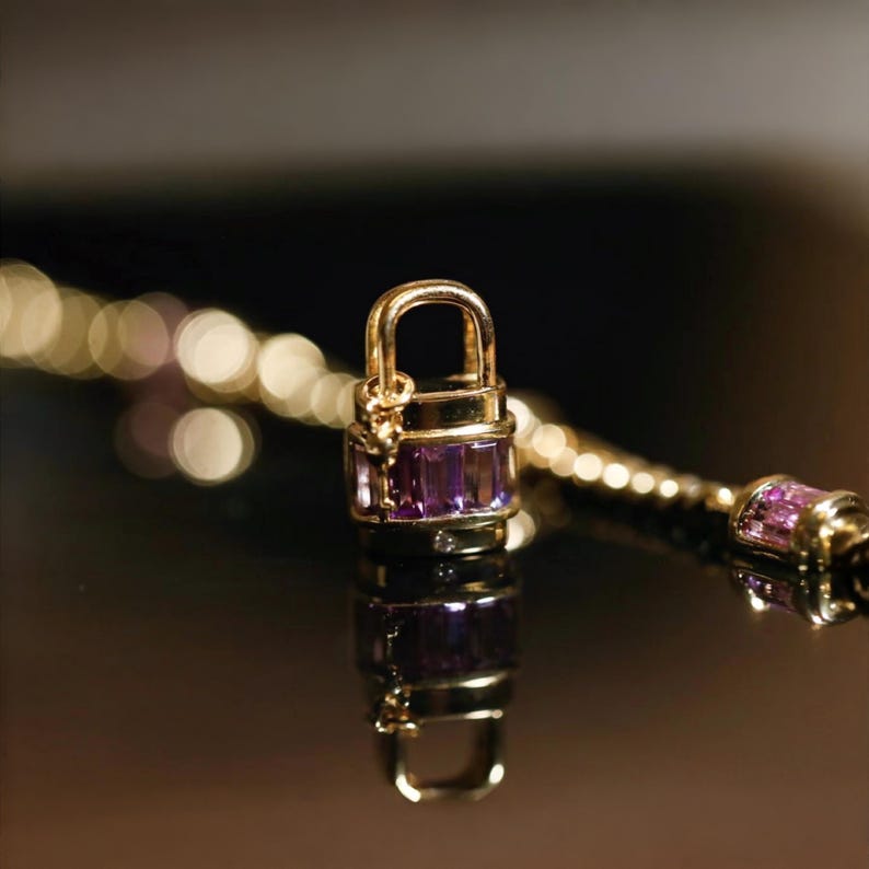 May include: A gold-colored padlock charm with a purple gemstone center, part of a necklace. The charm is rectangular with a curved handle. The necklace chain is also gold-colored, with a blurred background.