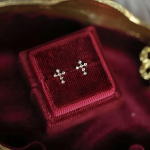 Może przedstawiać: Para małych złotych kolczyków w kształcie krzyża z małymi diamentami osadzonymi w kształcie krzyża. Kolczyki są prezentowane w czerwonym aksamitnym pudełku na biżuterię.