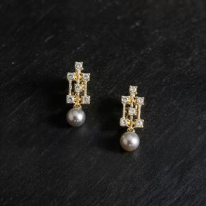 May include: A pair of gold earrings featuring a geometric, open-work rectangular frame set with small clear stones. Each earring is finished with a single, round, grey pearl drop hanging from the base of the metal structure.