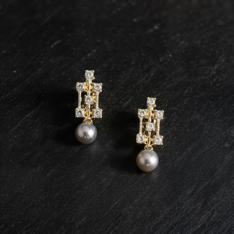 May include: A pair of gold earrings featuring a geometric, open-work rectangular frame set with small clear stones. Each earring is finished with a single, round, grey pearl drop hanging from the base of the metal structure.