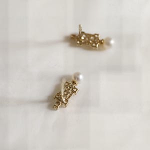 May include: A pair of gold-toned stud earrings featuring an intricate, openwork rectangular metal design. Each earring is accented with a single, small, round white pearl positioned at the top of the decorative metal structure.