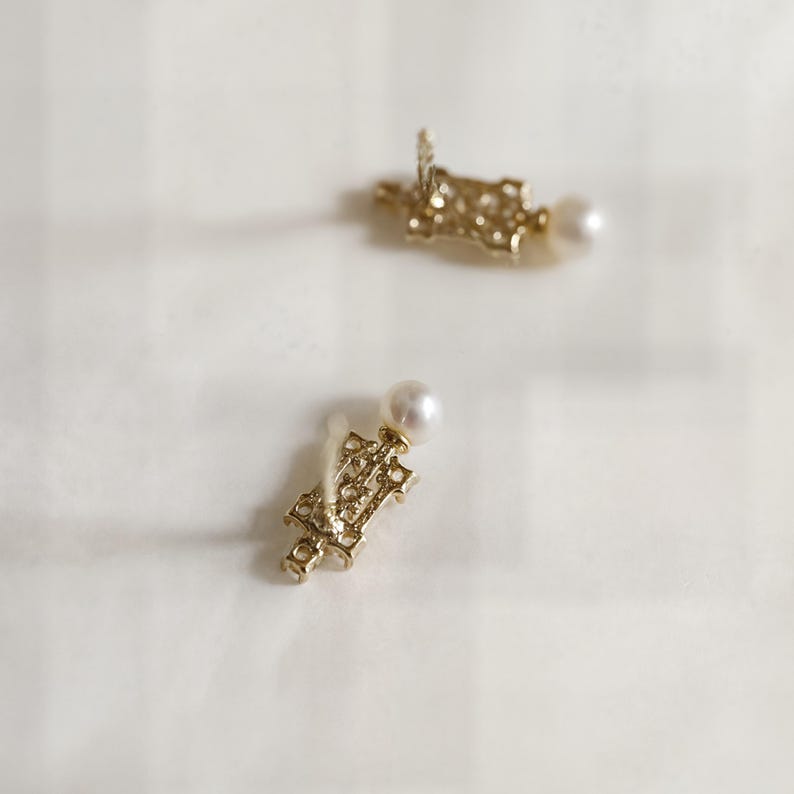 May include: A pair of gold-toned stud earrings featuring an intricate, openwork rectangular metal design. Each earring is accented with a single, small, round white pearl positioned at the top of the decorative metal structure.
