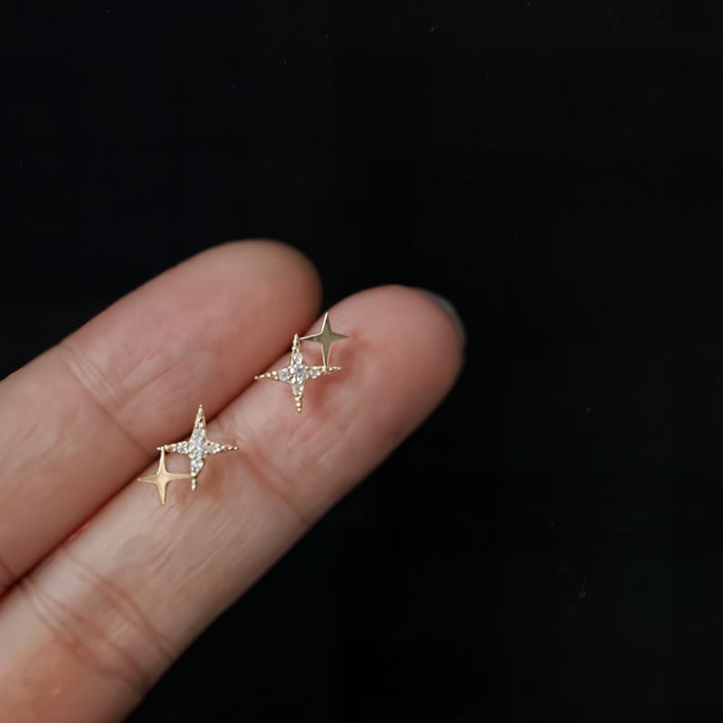 May include: A pair of gold-coloured star-shaped stud earrings. Each earring features a four-pointed star design, with one star adorned with small, clear gemstones. The earrings are displayed against a dark background.