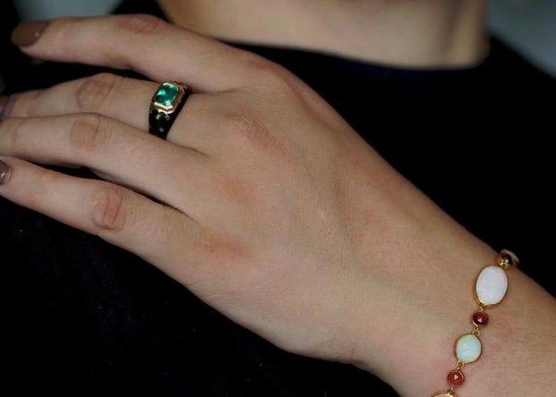 May include: A gold ring with a green gemstone set in a black enamel band. A gold bracelet with oval-shaped white opals and red gemstones.