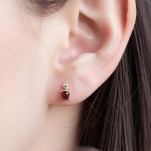 May include: A close-up of a gold stud earring with a square, deep red gemstone and a small, round, clear stone above it. The earring is set against the backdrop of a person's ear and hair.