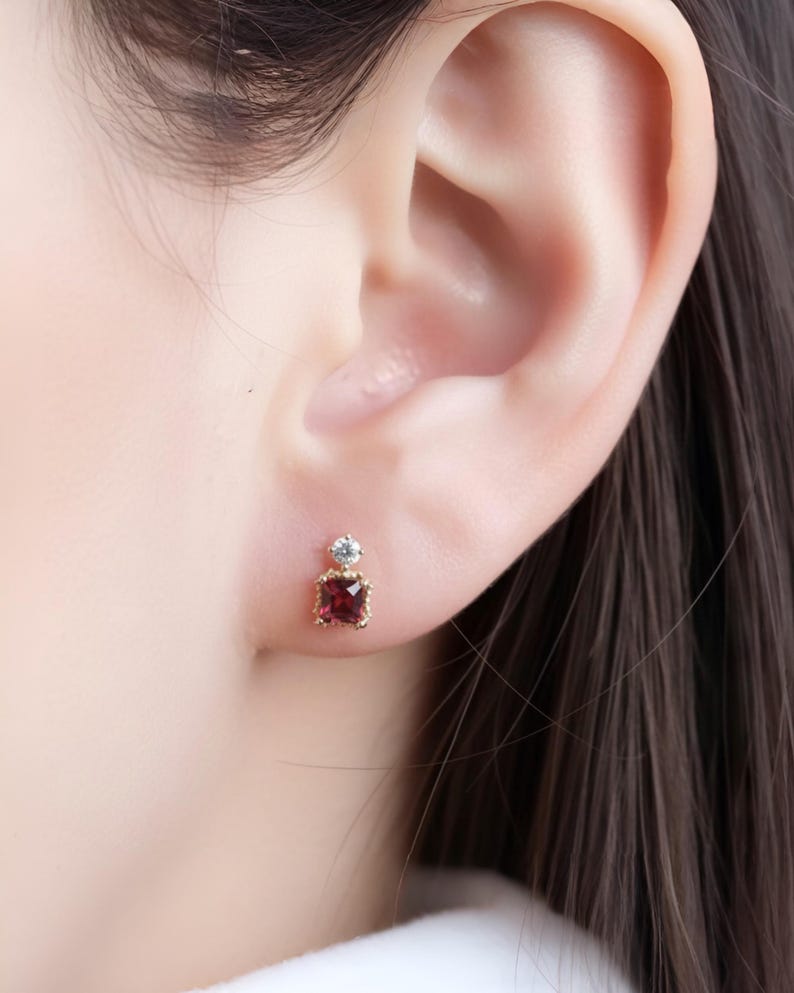May include: A close-up of a gold stud earring with a square, deep red gemstone and a small, round, clear stone above it. The earring is set against the backdrop of a person's ear and hair.