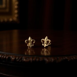 K&ouml;nnte beinhalten: Ein Paar goldfarbene Fleur-de-Lis-Ohrstecker. Die Ohrringe sind auf einer dunklen Holzoberfl&auml;che mit unscharfem Hintergrund platziert. Die Ohrringe sind klein und detailliert, mit einem klassischen Design.