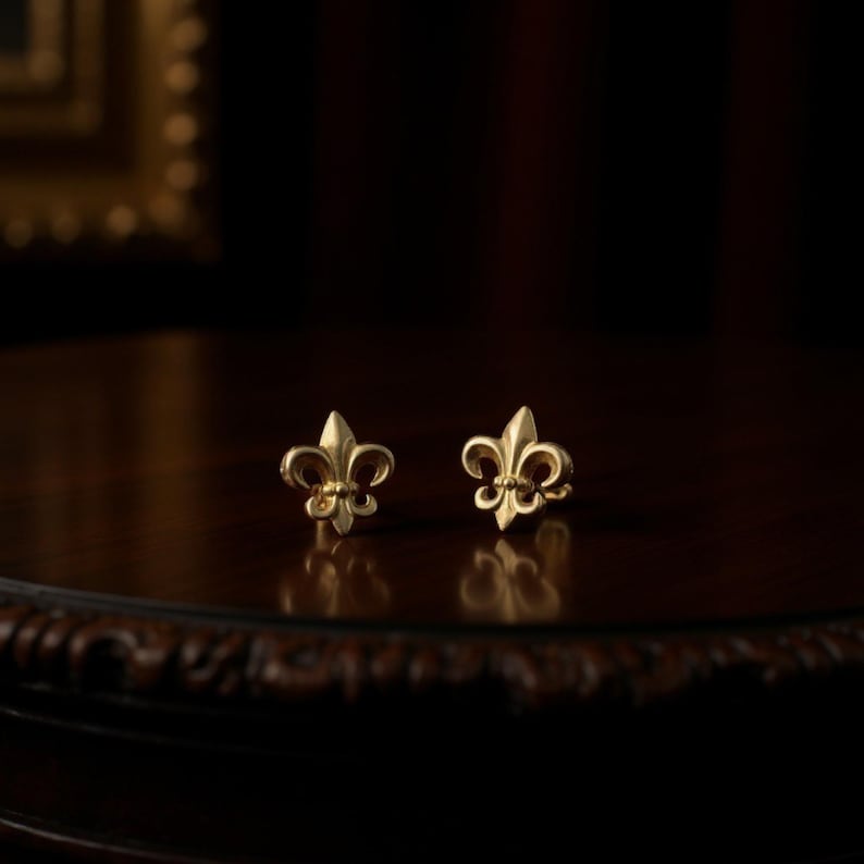 K&ouml;nnte beinhalten: Ein Paar goldfarbene Fleur-de-Lis-Ohrstecker. Die Ohrringe sind auf einer dunklen Holzoberfl&auml;che mit unscharfem Hintergrund platziert. Die Ohrringe sind klein und detailliert, mit einem klassischen Design.