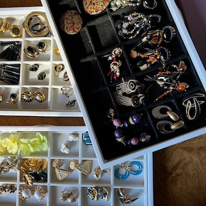 Op de afbeelding: Een witte sieradenorganisator met drie lagen, elk met een verscheidenheid aan oorbellen. De bovenste laag heeft zwarte vakken en bevat een mix van gouden, zilveren en kleurrijke oorbellen. De middelste laag heeft zwarte vakken en bevat een mix van gouden, zilveren en kleurrijke oorbellen. De onderste laag heeft witte vakken en bevat een mix van gouden, zilveren en kleurrijke oorbellen.