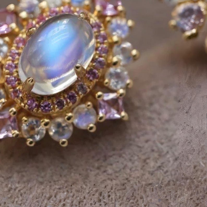 Puede incluir: Primer plano de una joya dorada con una gran piedra lunar ovalada e iridiscente. La piedra lunar est&aacute; rodeada de piedras preciosas rosas y transparentes m&aacute;s peque&ntilde;as, creando un efecto de halo. El engaste es detallado y ornamentado.