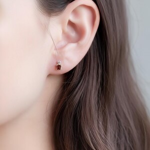 May include: Close-up of a small, square-cut, dark red gemstone earring with a small, clear stone above it. The earring is set in a gold-colored metal and is worn on an ear. The earring is a simple, elegant design.