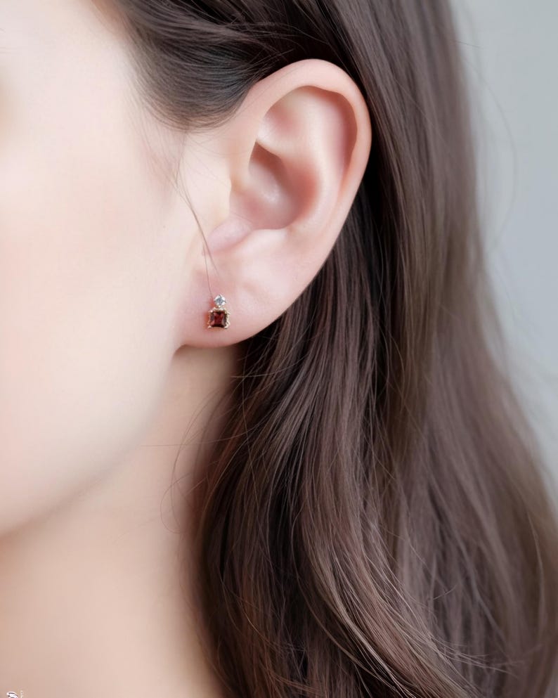 May include: Close-up of a small, square-cut, dark red gemstone earring with a small, clear stone above it. The earring is set in a gold-colored metal and is worn on an ear. The earring is a simple, elegant design.