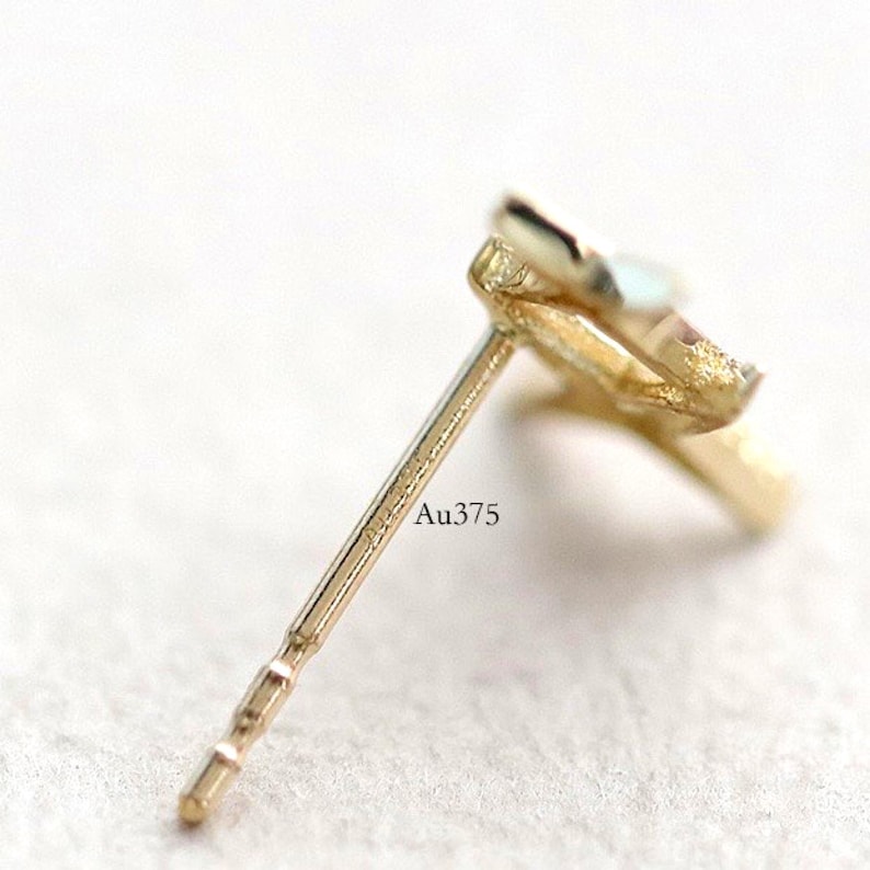 May include: Close-up of a gold stud earring. The earring post is visible, with the text "Au375" engraved on it. The earring has a delicate, possibly floral, design. The background is a soft, neutral colour.