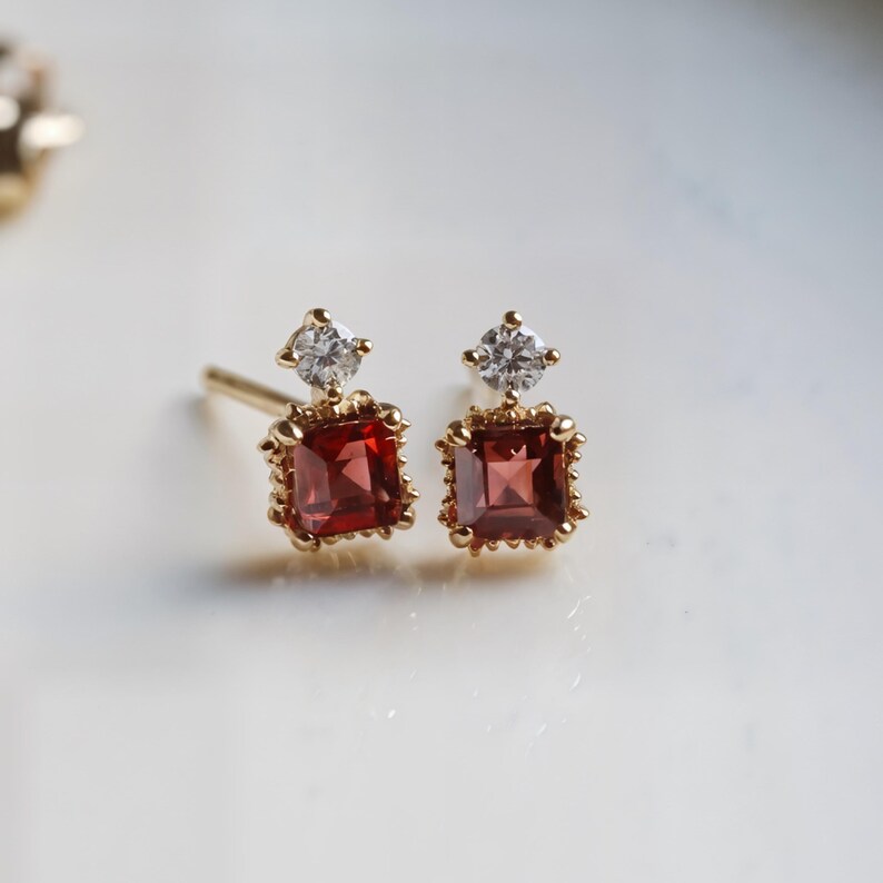 May include: A pair of gold stud earrings. Each earring features a square, deep red gemstone set in a gold frame, topped with a small, round, clear gemstone. The earrings are displayed on a white surface.