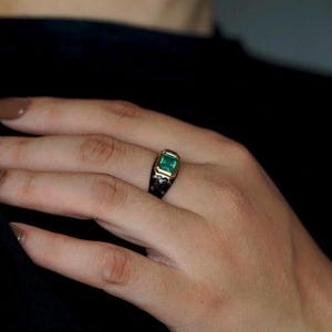 May include: A gold ring with a square emerald green gemstone set in a black enamel band.
