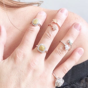 Puede incluir: Una mano con cuatro anillos. Dos de los anillos son de plata y presentan una piedra preciosa amarilla de forma cuadrada rodeada de piedras preciosas blancas m&aacute;s peque&ntilde;as. Los otros dos anillos son de oro y presentan un dise&ntilde;o de estrella.