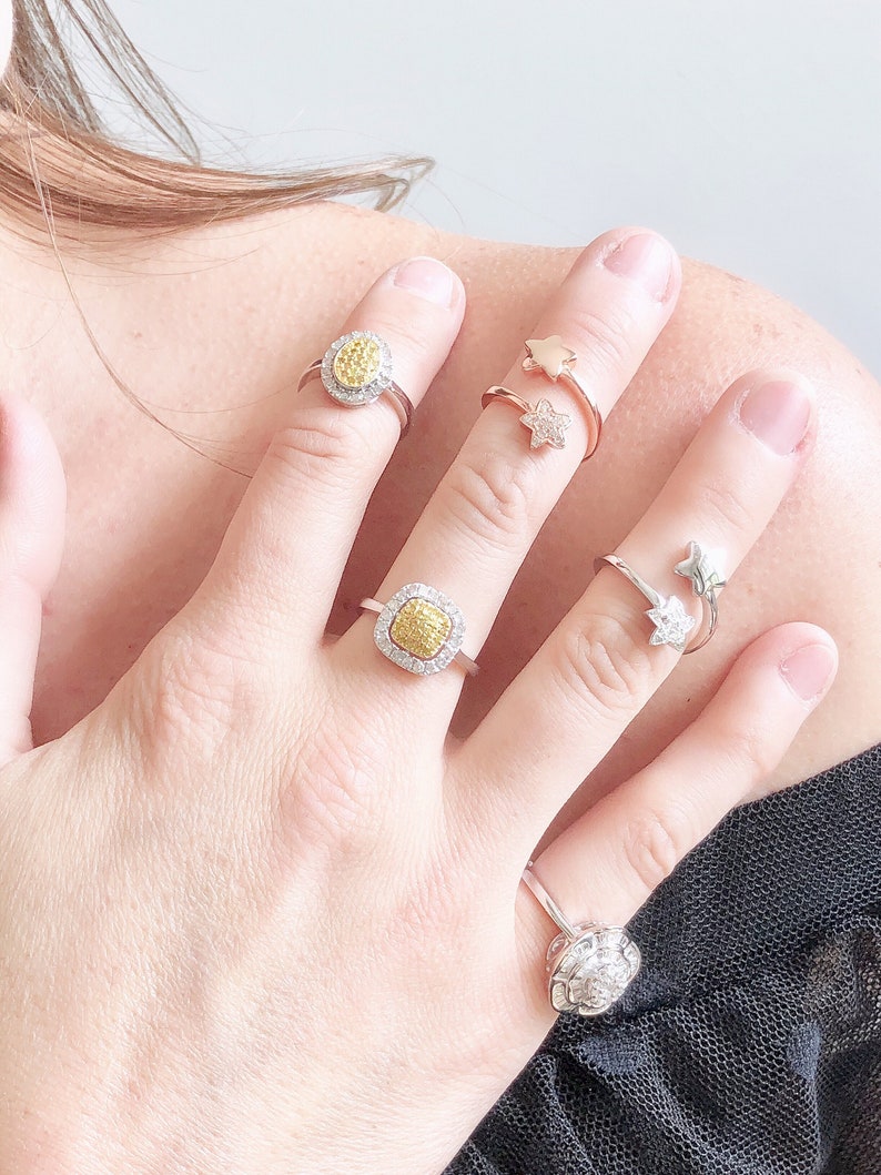 Puede incluir: Una mano con cuatro anillos. Dos de los anillos son de plata y presentan una piedra preciosa amarilla de forma cuadrada rodeada de piedras preciosas blancas m&aacute;s peque&ntilde;as. Los otros dos anillos son de oro y presentan un dise&ntilde;o de estrella.