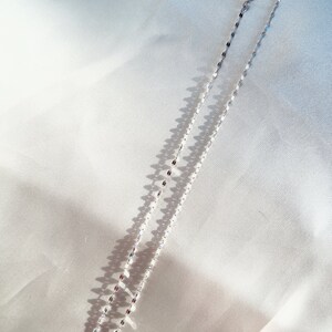May include: A delicate silver necklace with a repeating oval link design. The necklace is displayed on a white satin surface, with shadows adding depth. The chain is approximately 16 inches long.