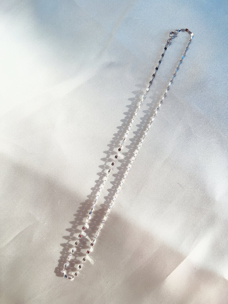 May include: A delicate silver necklace with a repeating oval link design. The necklace is displayed on a white satin surface, with shadows adding depth. The chain is approximately 16 inches long.