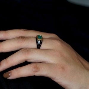 May include: A gold ring with a large emerald green gemstone set in a black setting. The ring is on a person's finger.