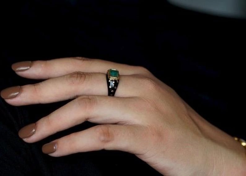 May include: A gold ring with a large emerald green gemstone set in a black setting. The ring is on a person's finger.