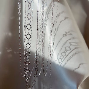 May include: A collection of delicate silver necklaces with various chain designs. The necklaces include chains with small beads, diamond-shaped links, and wavy patterns. The jewelry is displayed against a white, textured background, with sunlight casting shadows.