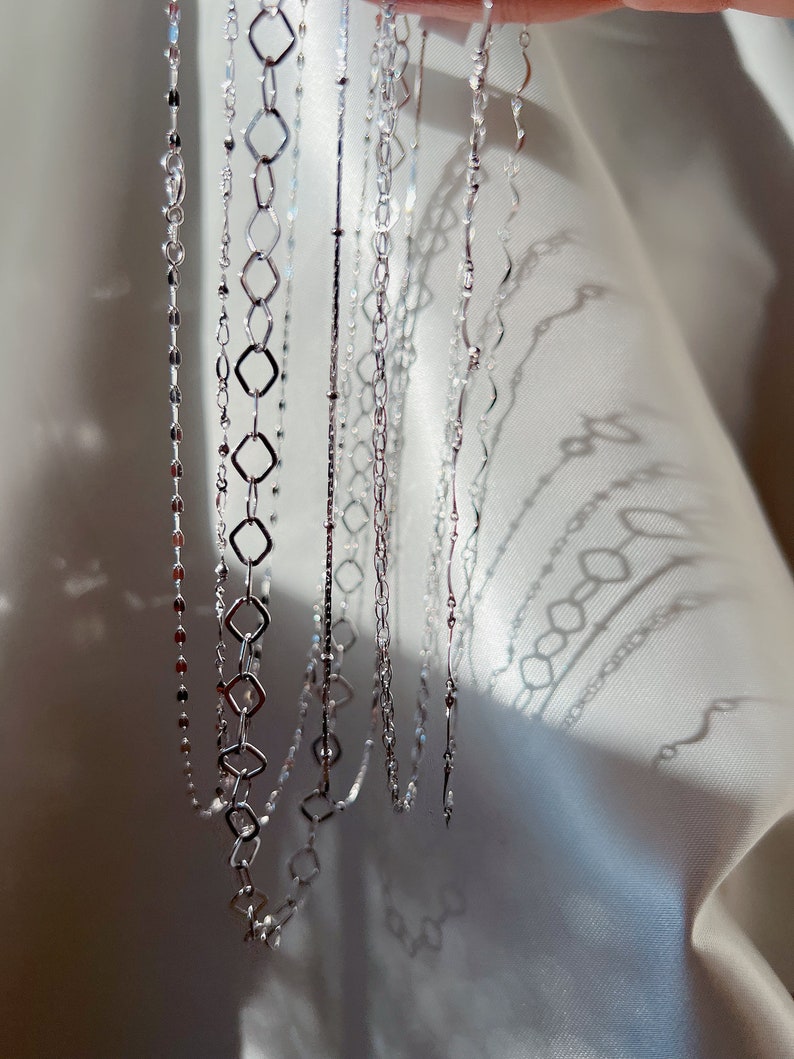 May include: A collection of delicate silver necklaces with various chain designs. The necklaces include chains with small beads, diamond-shaped links, and wavy patterns. The jewelry is displayed against a white, textured background, with sunlight casting shadows.