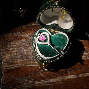 May include: A heart-shaped jewelry box with a green velvet interior. A silver key charm with a pink gemstone is inside the box.