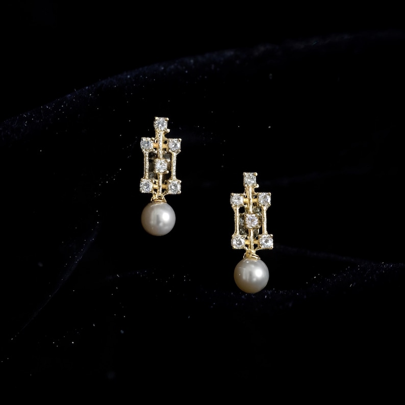 May include: A pair of gold earrings featuring a rectangular openwork frame set with small clear stones. Each earring is accented with a single round white pearl dangling from the base of the geometric metal structure.