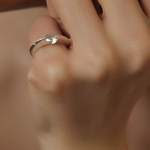 May include: A silver ring with a single, clear, round gemstone set in the center. The ring is worn on a finger.