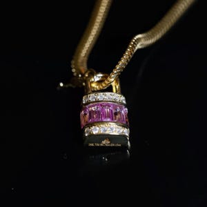 May include: A gold necklace with a pendant shaped like a padlock. The padlock is adorned with pink and clear gemstones. The necklace is on a black background. The pendant has the text "18K 750 SC 0.78ct Do.13ct."