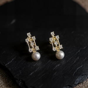 May include: A pair of gold-tone stud earrings featuring a rectangular openwork frame set with small clear stones and a single round white pearl dangling from the base of each piece, creating a balanced and symmetrical jewelry design.