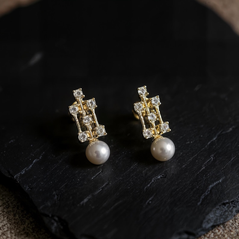 May include: A pair of gold-tone stud earrings featuring a rectangular openwork frame set with small clear stones and a single round white pearl dangling from the base of each piece, creating a balanced and symmetrical jewelry design.