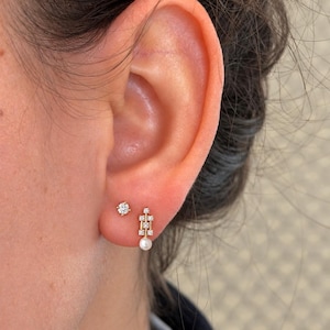 May include: A gold stud earring featuring a single round clear stone and a gold drop earring with a vertical bar design, three small clear stones, and a small white pearl at the base, both worn in the earlobe.