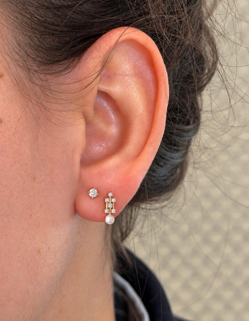 May include: A gold stud earring featuring a single round clear stone and a gold drop earring with a vertical bar design, three small clear stones, and a small white pearl at the base, both worn in the earlobe.