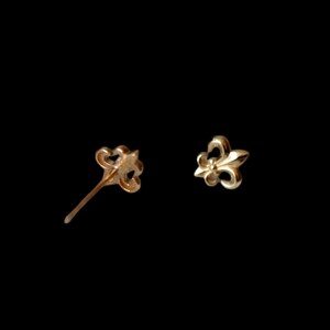 K&ouml;nnte beinhalten: Zwei goldfarbene Fleur-de-Lis-Ohrstecker. Ein Ohrring ist ein Stecker, der andere ein Pfosten. Die Ohrringe sind vor einem schwarzen Hintergrund platziert. Die Ohrringe sind klein und zart.