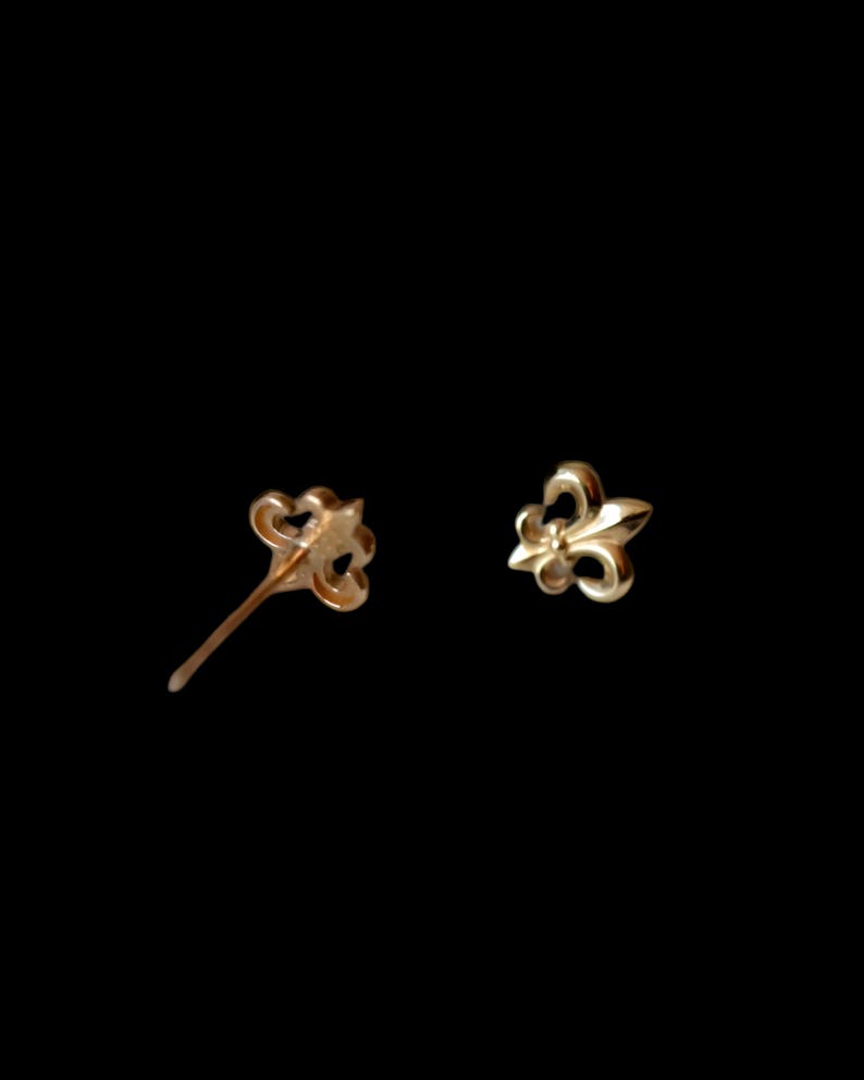 K&ouml;nnte beinhalten: Zwei goldfarbene Fleur-de-Lis-Ohrstecker. Ein Ohrring ist ein Stecker, der andere ein Pfosten. Die Ohrringe sind vor einem schwarzen Hintergrund platziert. Die Ohrringe sind klein und zart.