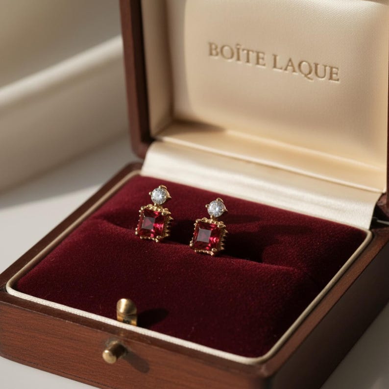 May include: A pair of gold earrings with square, red gemstones and a small, clear stone on top, displayed in a wooden box with a red velvet interior. The box is open, revealing the earrings and the text "BOITE LAQUE".