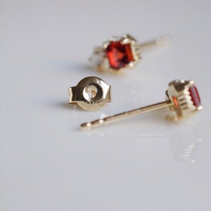 May include: Close-up of gold stud earrings with a vibrant red gemstone. The earrings have a classic design with a round gemstone setting. The earring backs are also visible, showing the post and clasp mechanism. The earrings are set against a white background.