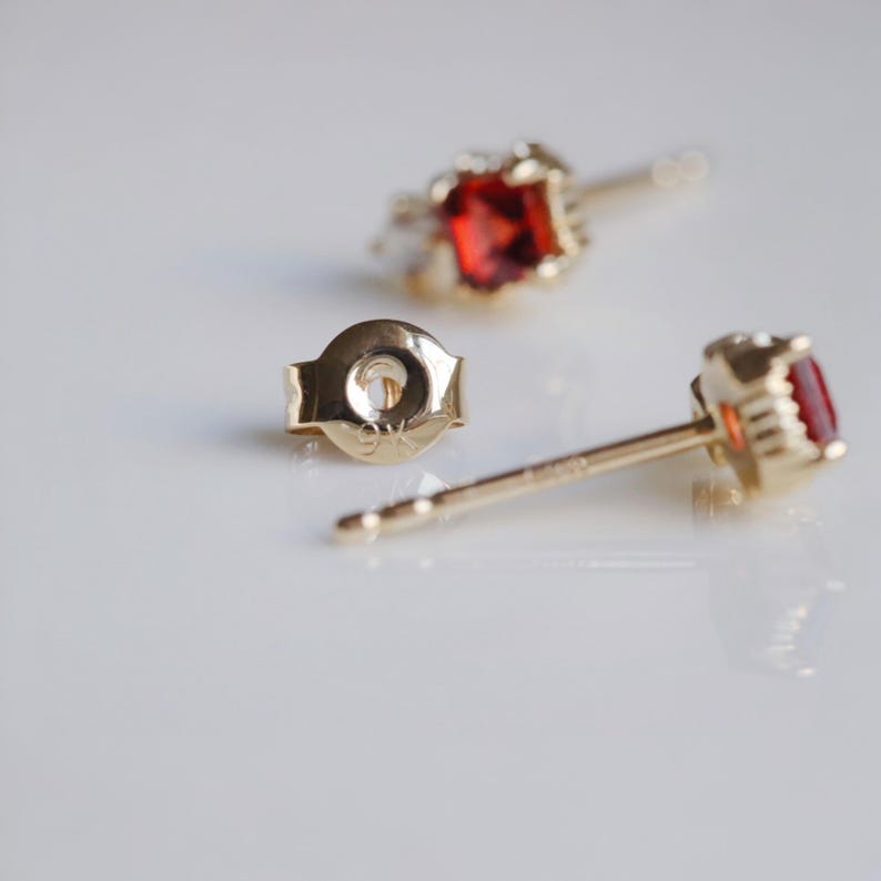 May include: Close-up of gold stud earrings with a vibrant red gemstone. The earrings have a classic design with a round gemstone setting. The earring backs are also visible, showing the post and clasp mechanism. The earrings are set against a white background.