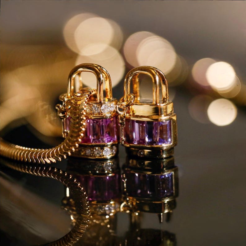 May include: Two gold-colored padlock pendants with purple and clear gemstones. The pendants are connected to a gold chain. The background is blurred with warm, golden tones, creating a luxurious feel. The pendants are reflected on a dark surface.