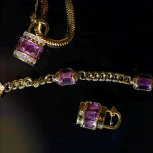 May include: A gold-toned jewelry set featuring a necklace with a padlock pendant, a bracelet, and a padlock-shaped earring. The pendants and bracelet links are adorned with rectangular pink gemstones and small white diamonds. The jewelry is set against a black background.
