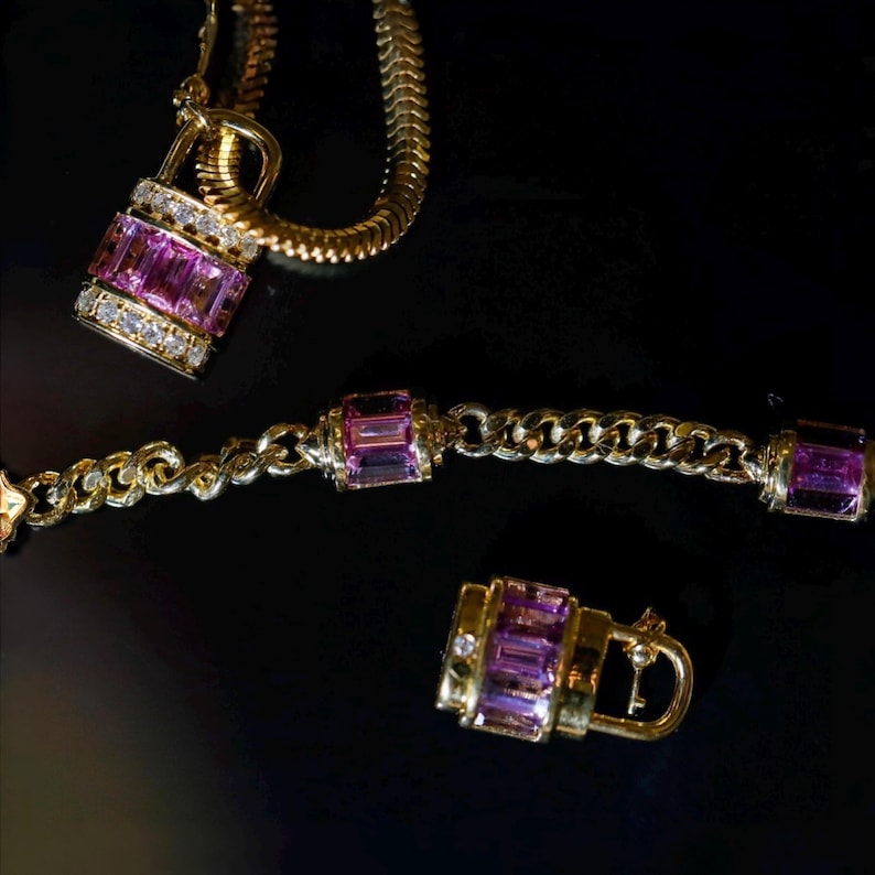 May include: A gold-toned jewelry set featuring a necklace with a padlock pendant, a bracelet, and a padlock-shaped earring. The pendants and bracelet links are adorned with rectangular pink gemstones and small white diamonds. The jewelry is set against a black background.