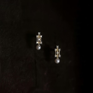 May include: A pair of gold drop earrings featuring a vertical rectangular frame set with small clear stones and a single round white pearl dangling from the base of each piece, presented against a dark, solid background.