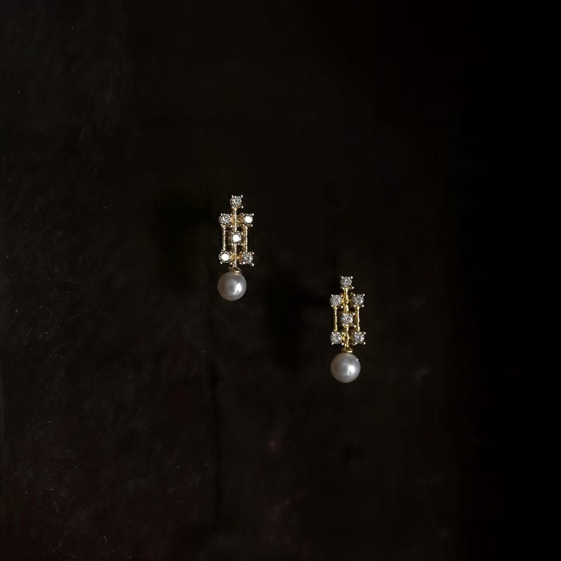 May include: A pair of gold drop earrings featuring a vertical rectangular frame set with small clear stones and a single round white pearl dangling from the base of each piece, presented against a dark, solid background.