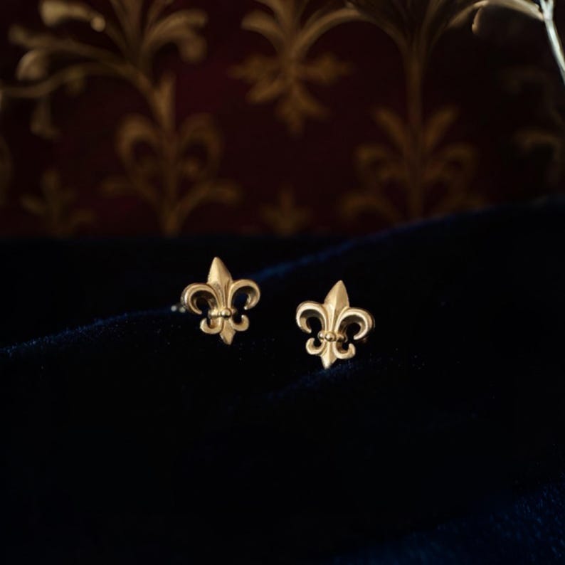 K&ouml;nnte beinhalten: Ein Paar goldfarbene Fleur-de-Lis-Ohrstecker. Die Ohrringe werden auf einer dunkelblauen Samtoberfl&auml;che mit einem gemusterten bordeauxfarbenen Hintergrund pr&auml;sentiert. Die Ohrringe sind klein und detailliert, mit einem klassischen Design.