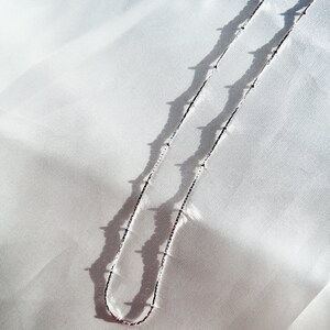 May include: A delicate silver necklace with small, evenly spaced beads. The necklace is displayed on a white, textured fabric, with shadows creating a contrasting effect. The chain is thin and elegant, suitable for everyday wear.